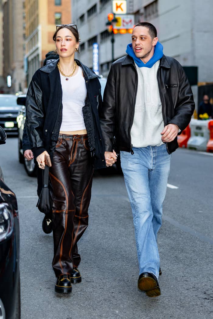 Two people walk hand in hand on a city street. One wears a casual jacket with jeans, and the other sports a stylish outfit with a white top and leather pants