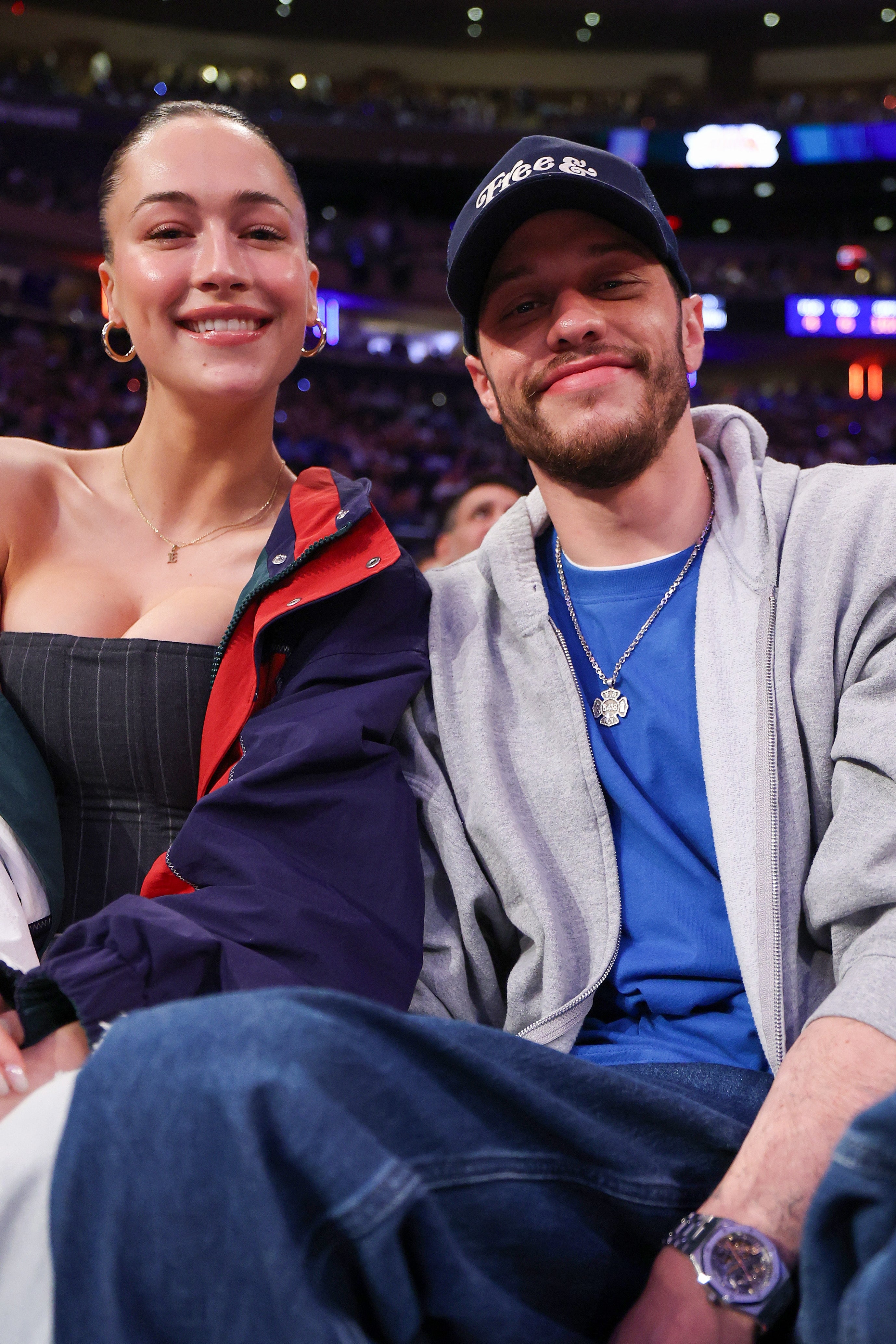 Two people smiling at a sports event, seated closely. One wears a strapless dress and hoop earrings. The other wears a baseball cap, necklace, and casual jacket