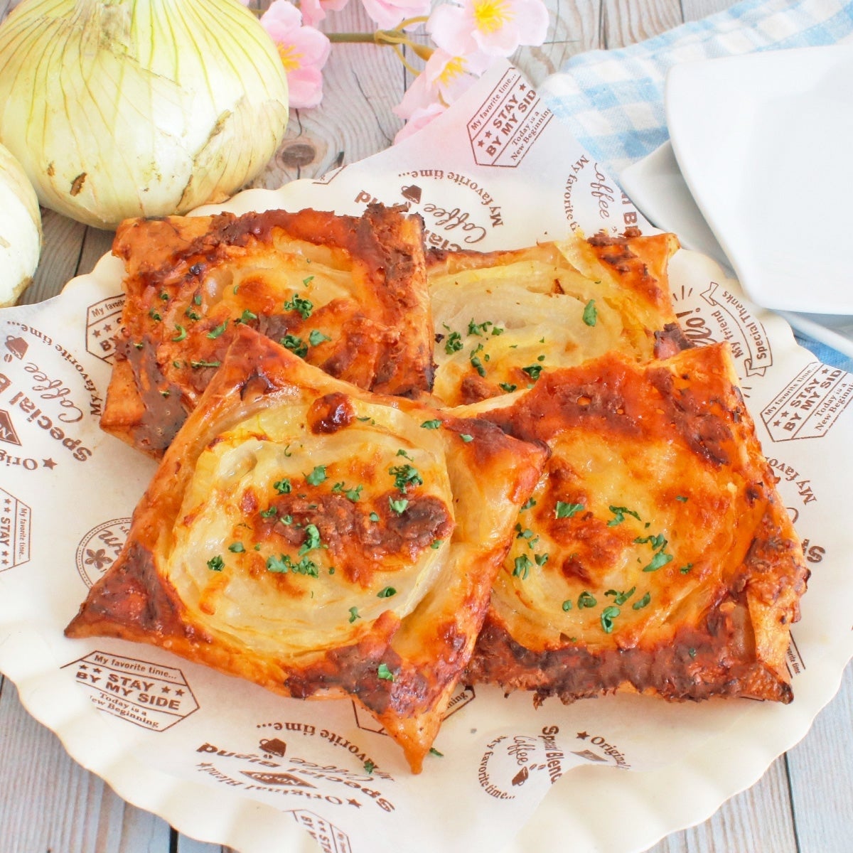 玉ねぎとチーズがトッピングされた四つのパイが皿に盛られている。周囲に玉ねぎと桜の花が飾られている。