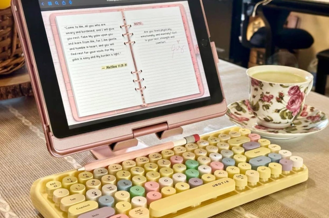 Tablet on a stand displays a digital journal next to a colorful retro-style keyboard and a floral teacup on a saucer