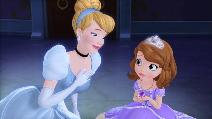 An animated Cinderella kneels beside Sofia, who is sitting down holding an amulet, both in elegant gowns and within a palace setting