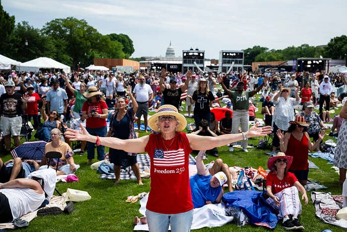 A large crowd at the Rededicate 250 event with the U.S. Capitol in the background. A woman amongst the crowd hold her arms out wide with passion