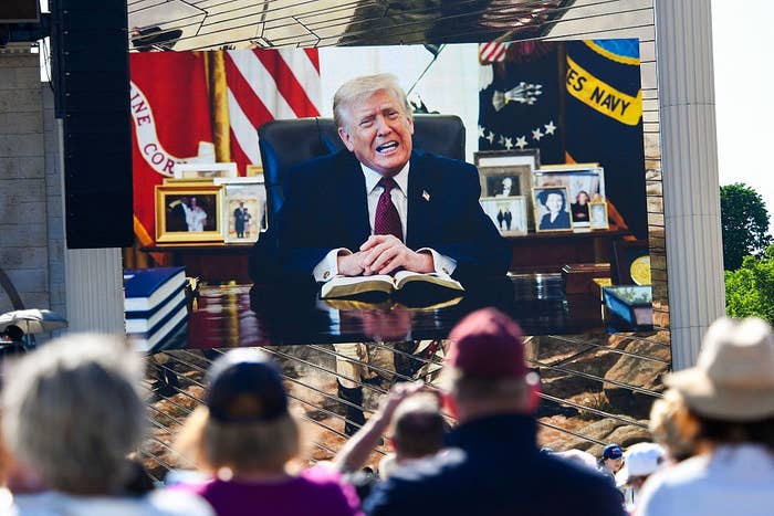 A crowd watches a large screen showing Trump reciting a Bible passage in the Oval Office