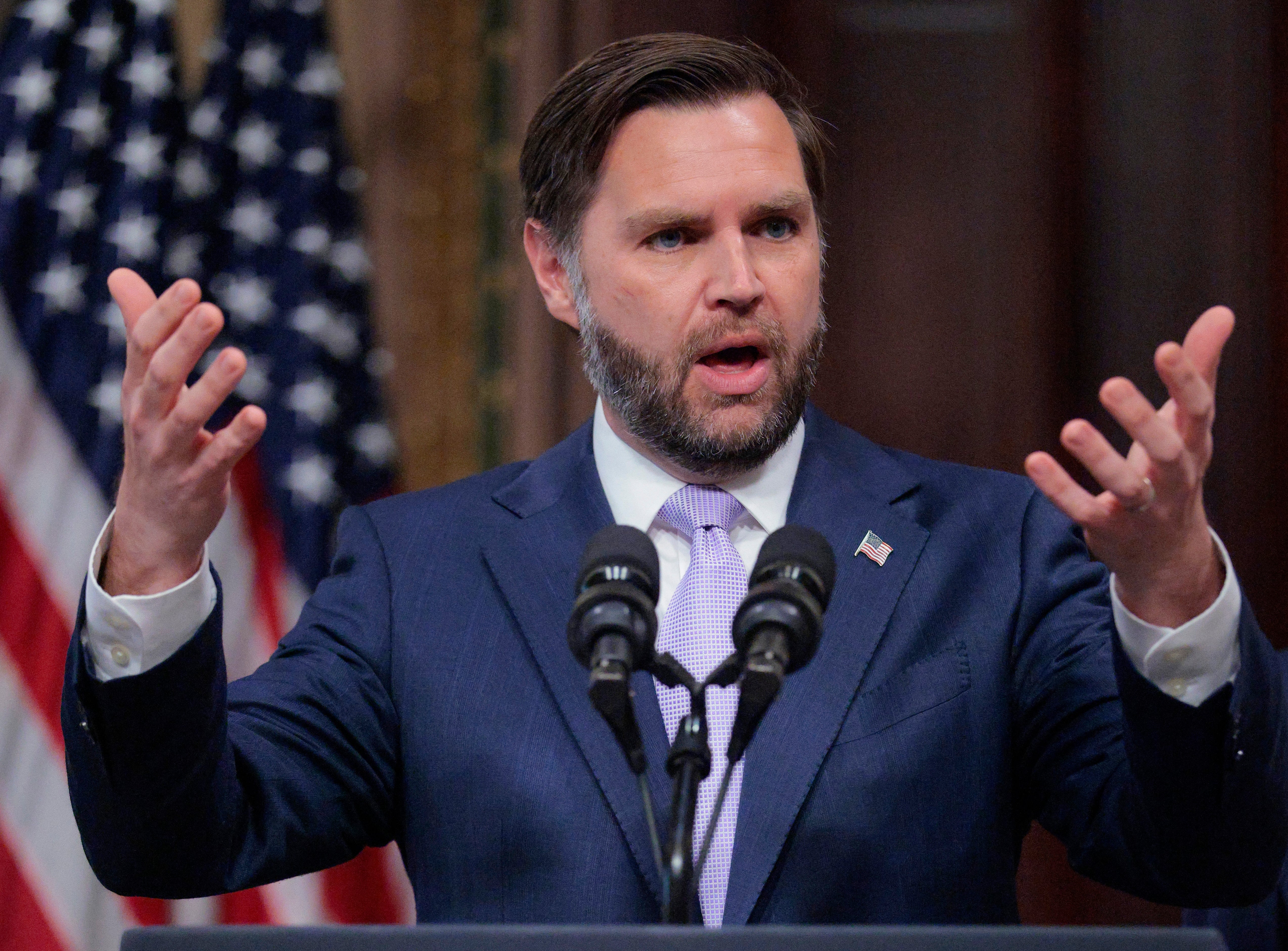 A man in a suit speaks passionately at a podium with microphones, a U.S. flag in the background