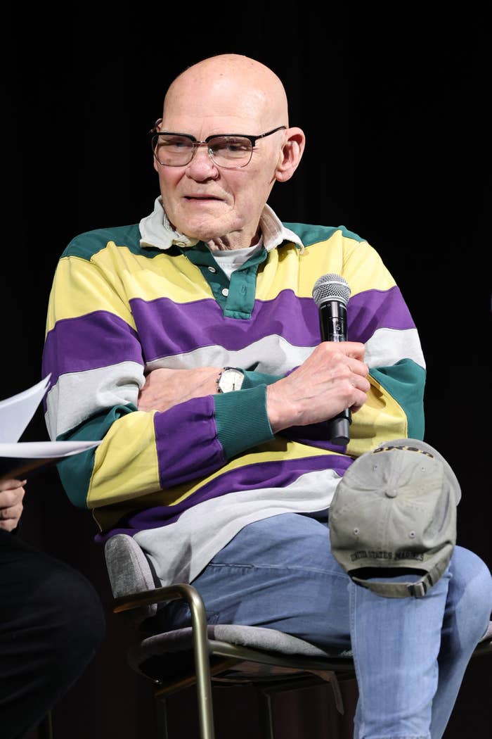 An older man with glasses and a striped rugby shirt holds a microphone while seated on stage, participating in a discussion