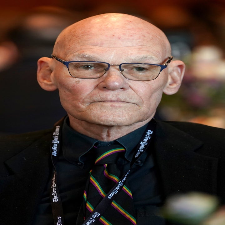 A person wearing glasses and a suit at a formal event, looking pensive, surrounded by other attendees in the background