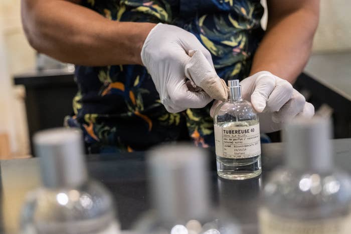 Person wearing gloves opens a vessel  labeled "Tubereuse 40" connected  a countertop with different   akin  bottles