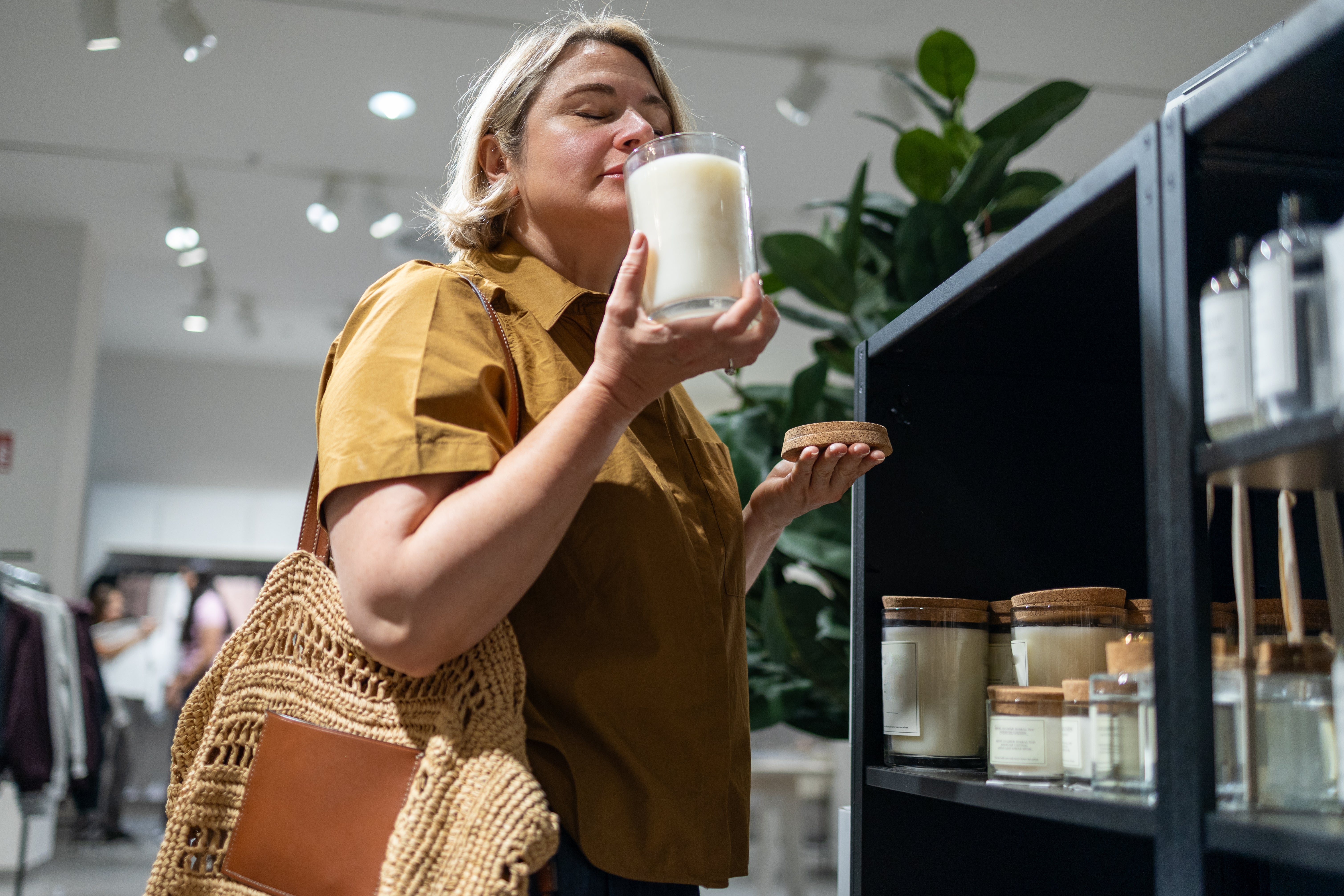 Person enjoying the scent of a candle successful a store, holding the lid successful 1 manus and carrying a woven container implicit their shoulder