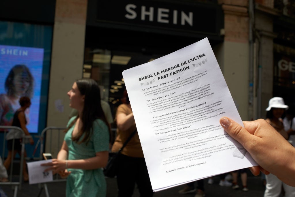 Person holds a document titled "SHEIN: La Marque de l'Ultra Fast Fashion" outside a SHEIN store, with people passing by