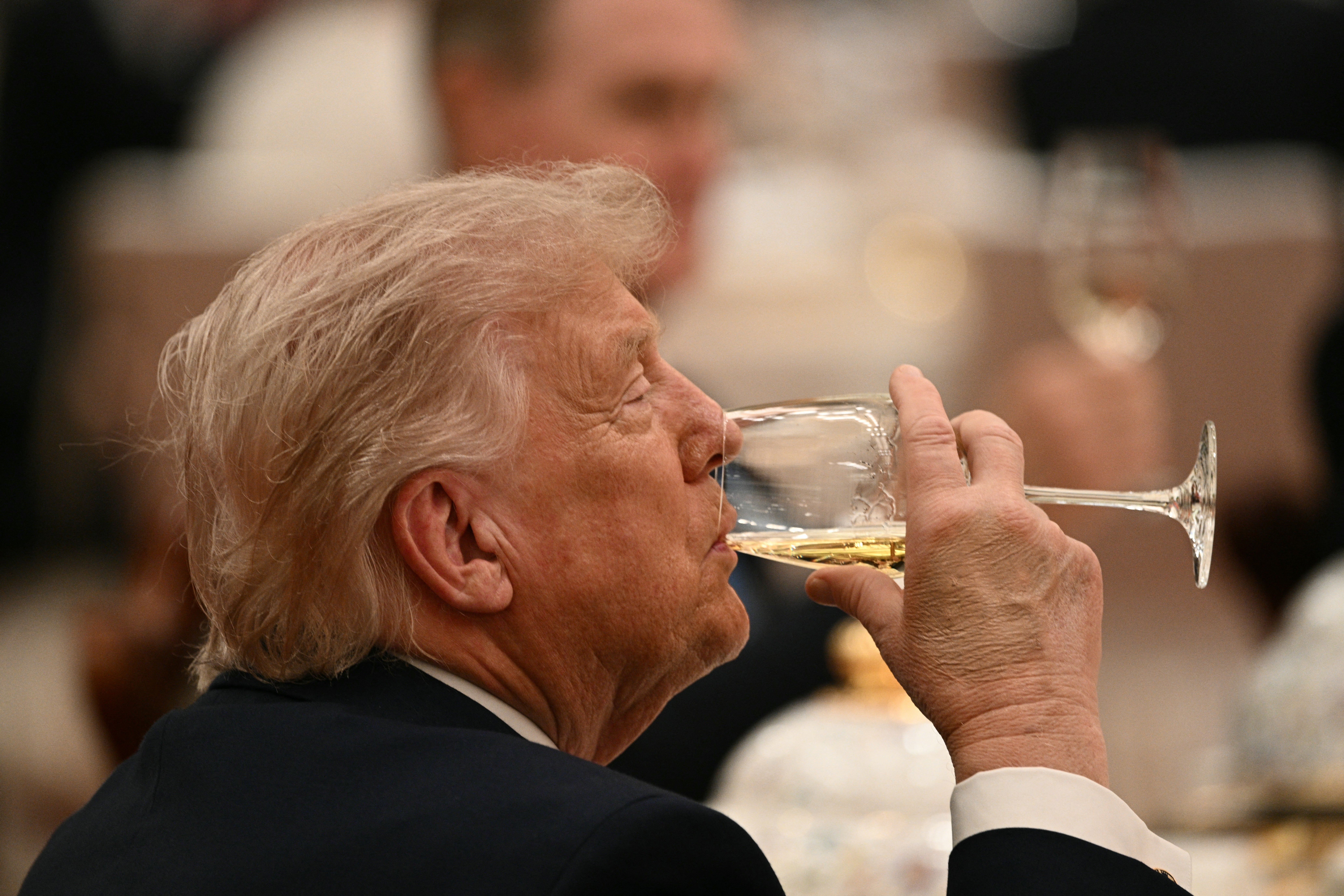 Person in formal attire sipping from a wine glass at an elegant event, other attendees in the background