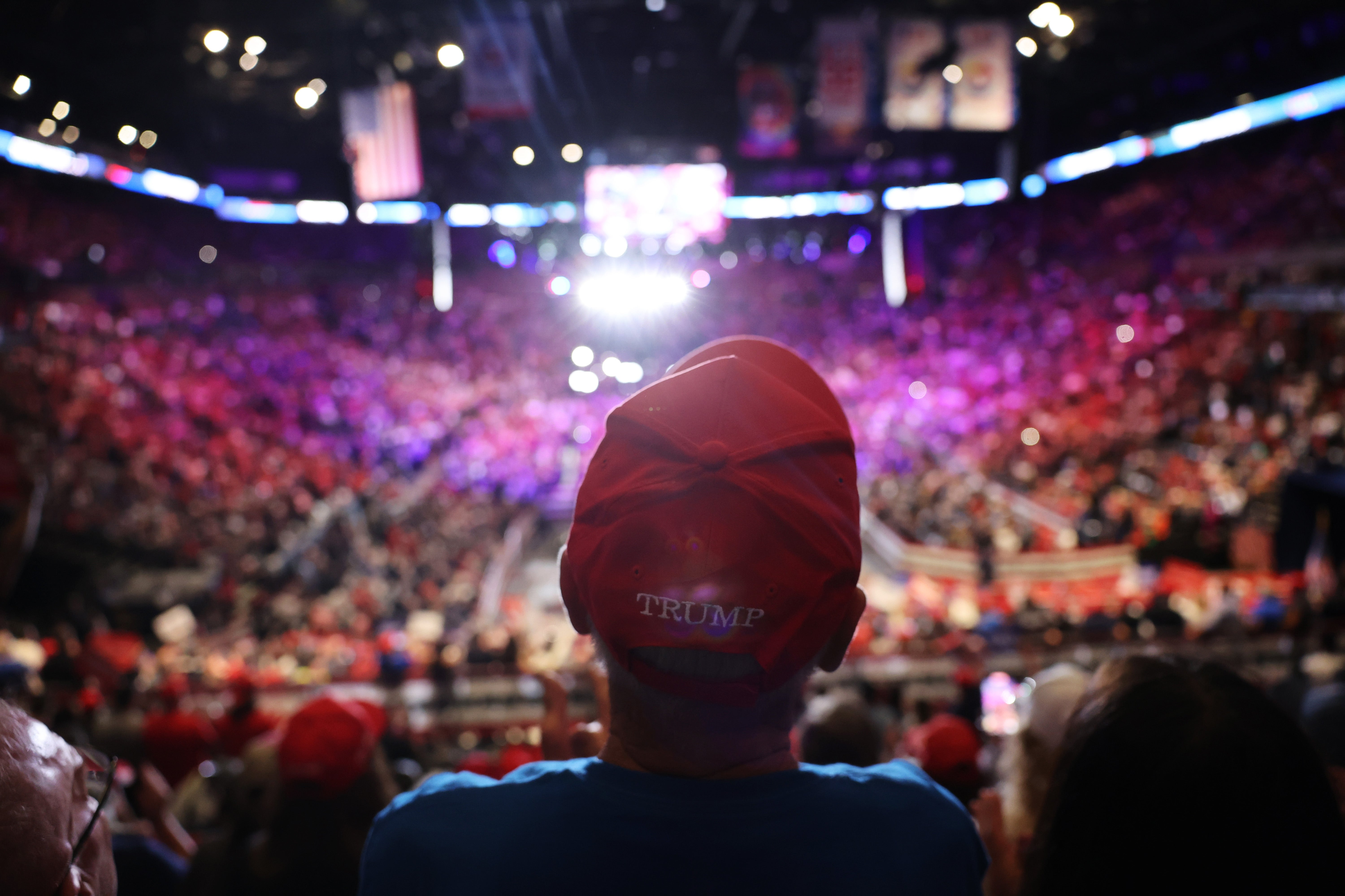 Person in a hat with "Trump" facing a crowded arena, vibrant lights and many people in the background