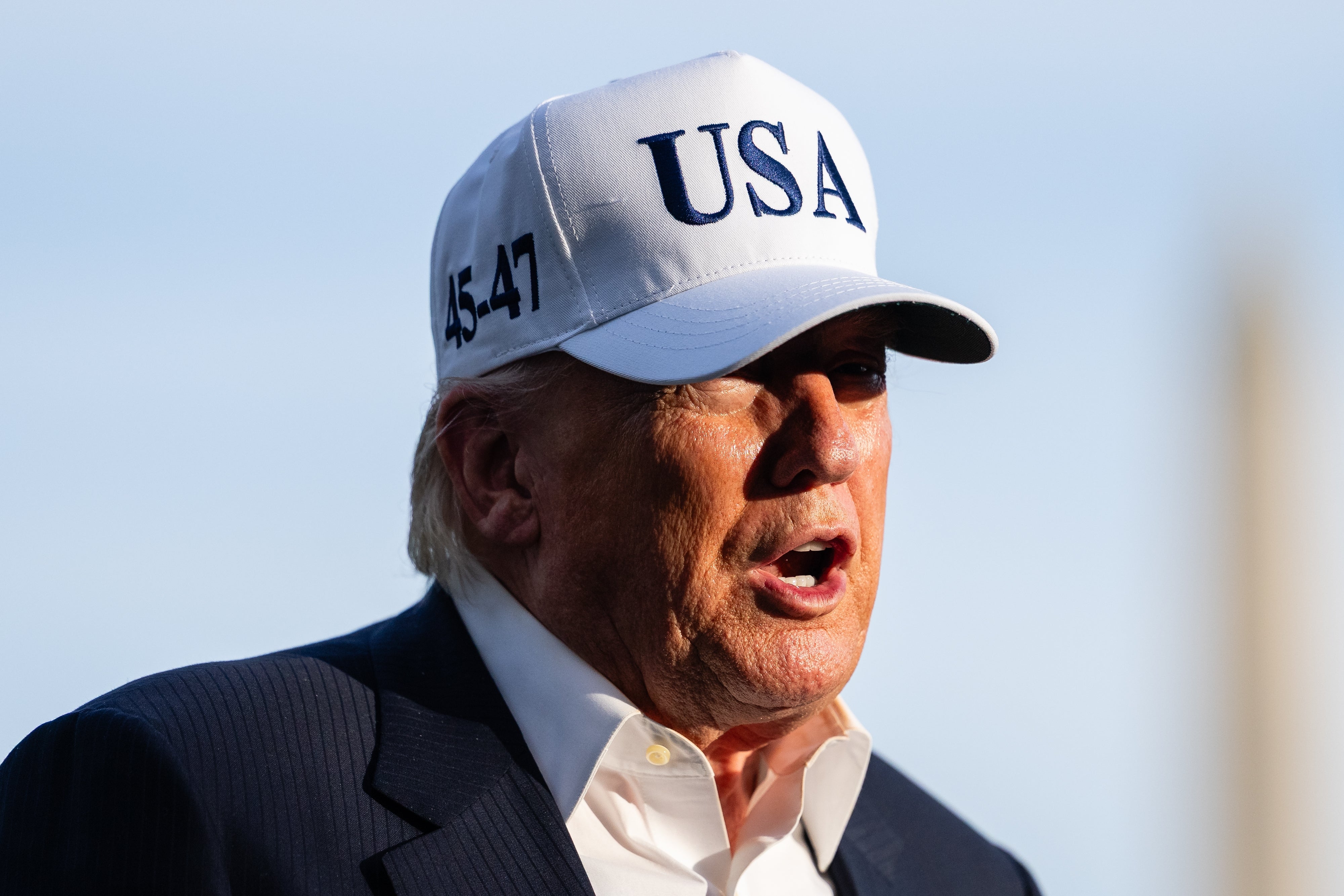Person wearing a "USA" cap with "45-47" on the side, in a suit, speaking outdoors