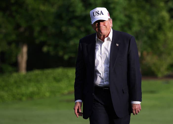 Man in a suit and "USA" cap walking outdoors on grass, smiling