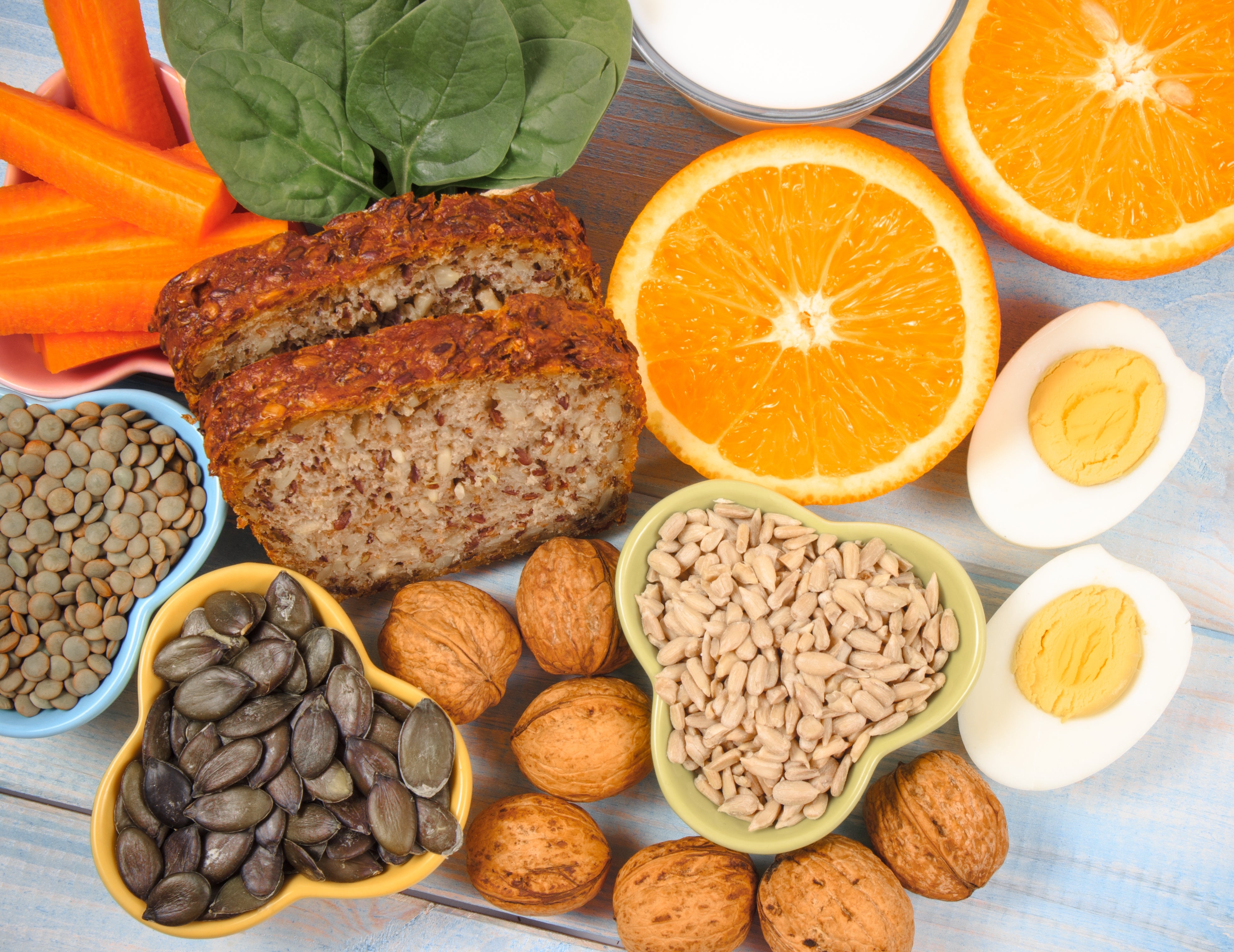 A assortment of steadfast foods including bread, oranges, boiled eggs, nuts, seeds, lentils, carrots, and spinach are arranged connected a table