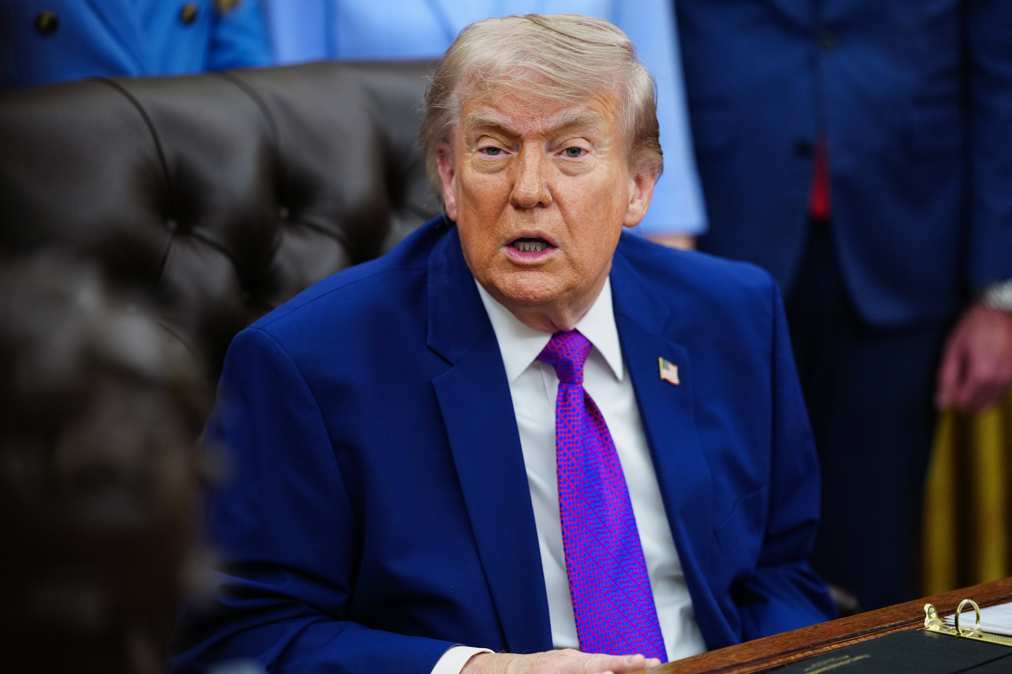 Person seated at a desk wearing a suit with a bright tie, speaking in a meeting setting with people nearby