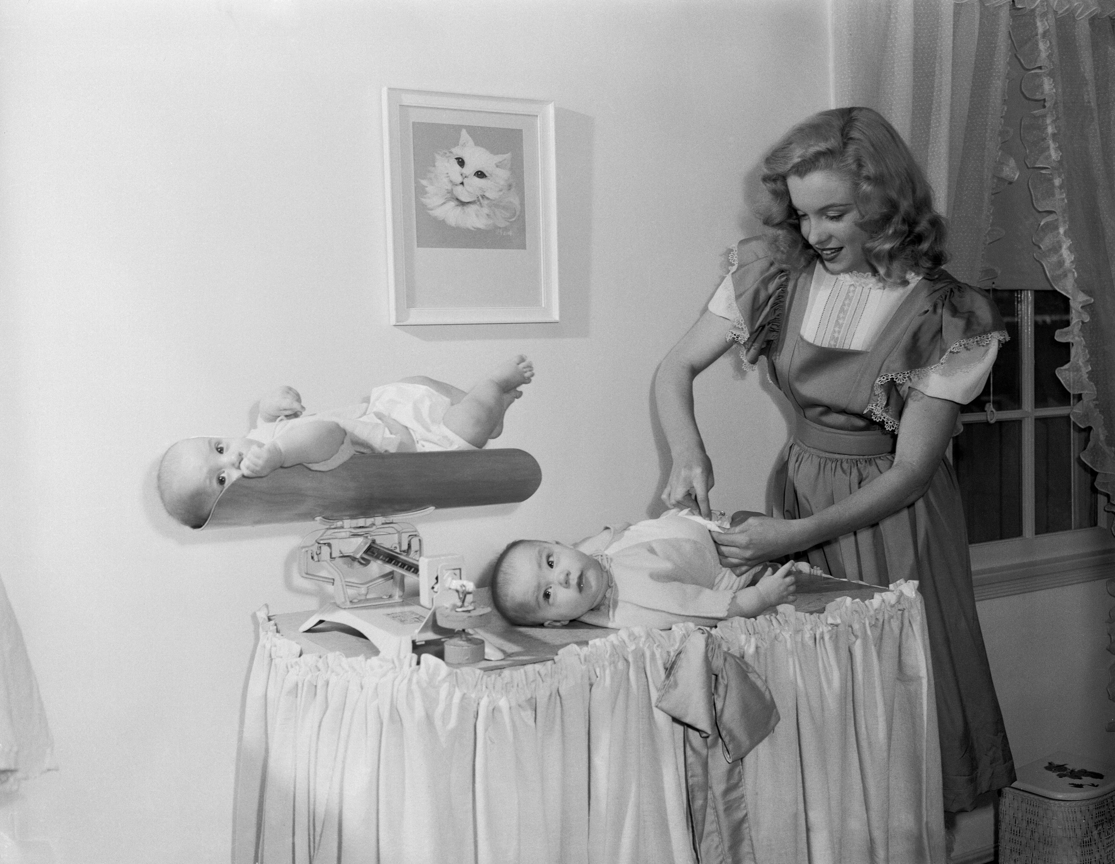 Marilyn Monroe changes a baby's diaper on a changing table, with another baby lying on a nearby shelf. A cat picture hangs on the wall