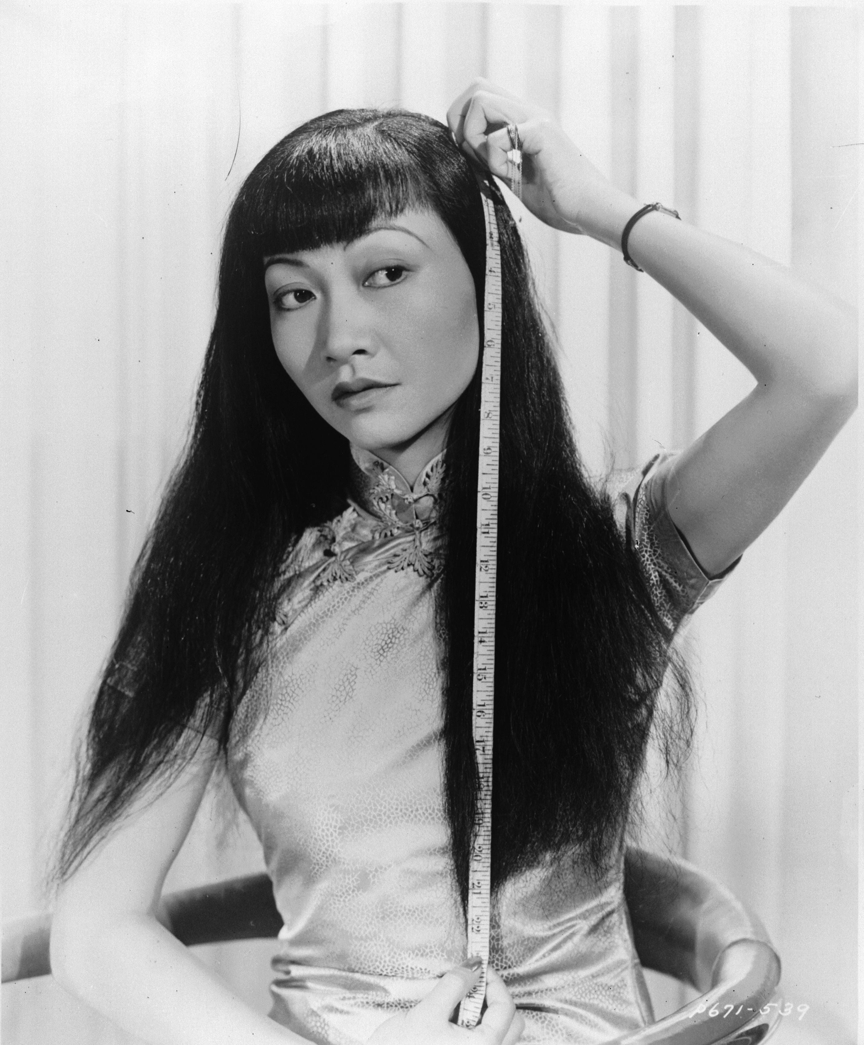 Anna May Wong in a traditional dress measures her long hair, looking contemplative