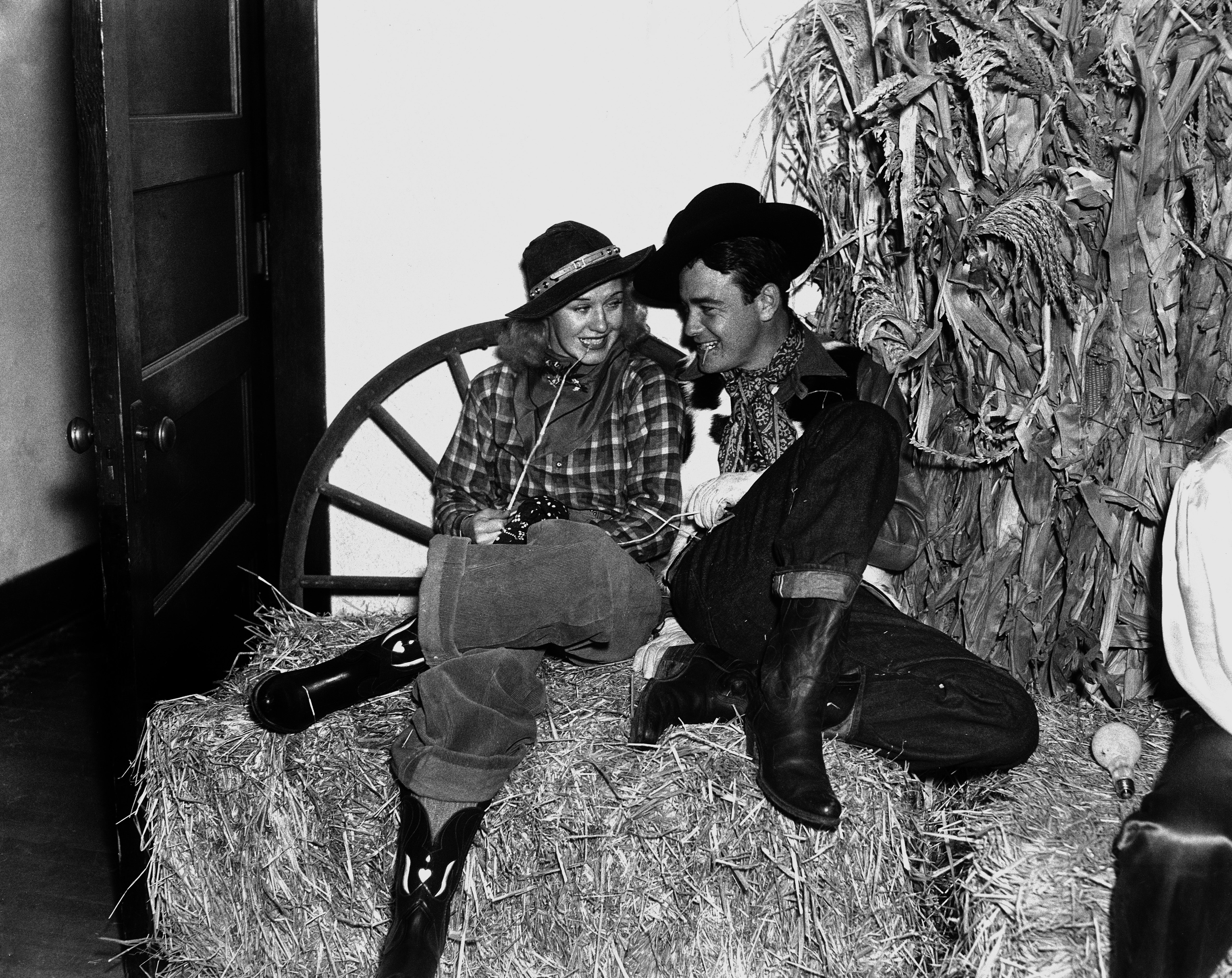 Ginger Rogers and Lew Ayres dressed as cowboys sit on hay bales, chatting happily next to a corn stalk display