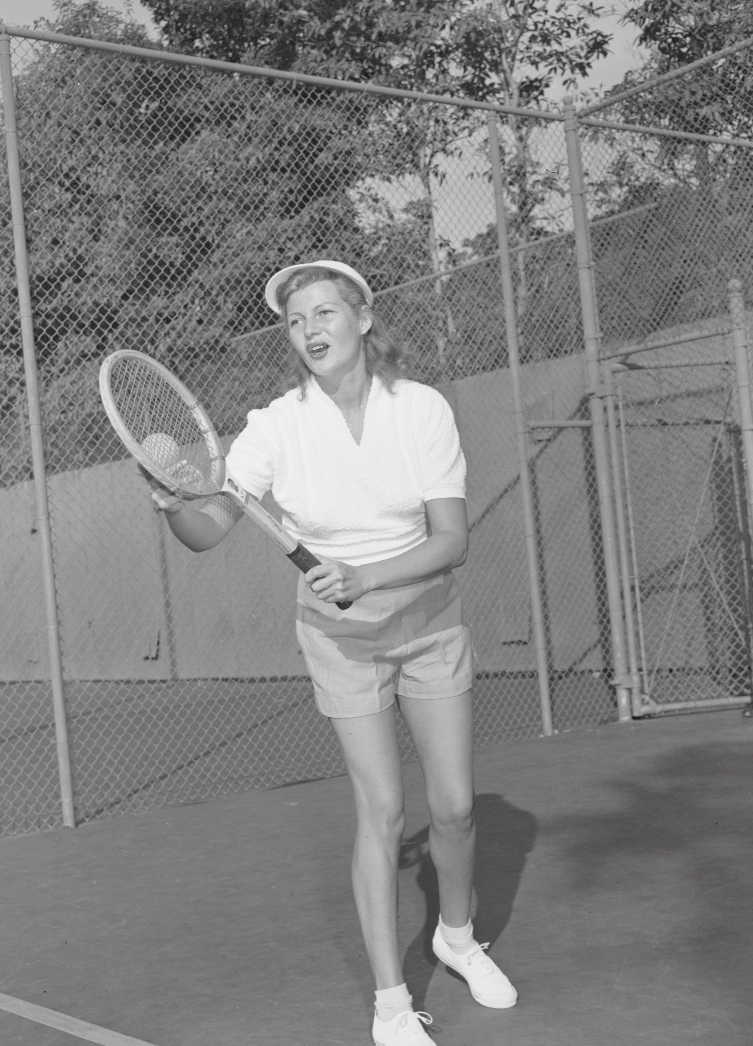 Rita Hayworth playing tennis on a court, wearing a shorts and a polo shirt, with a visor, holding a tennis racket, ready to hit a ball
