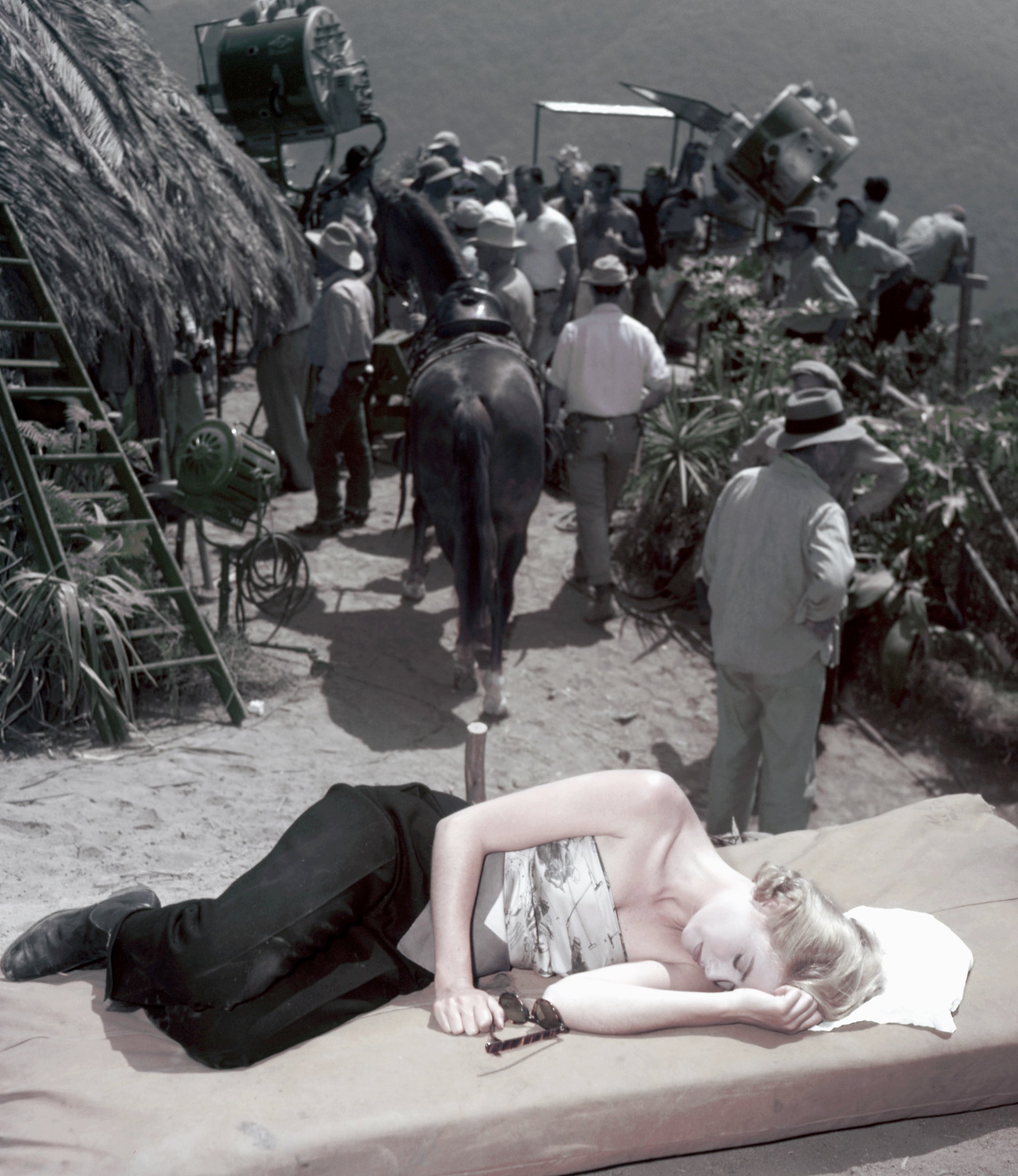 Grace Kelly resting on a mattress on a film set with crew, cameras, and a horse in the background