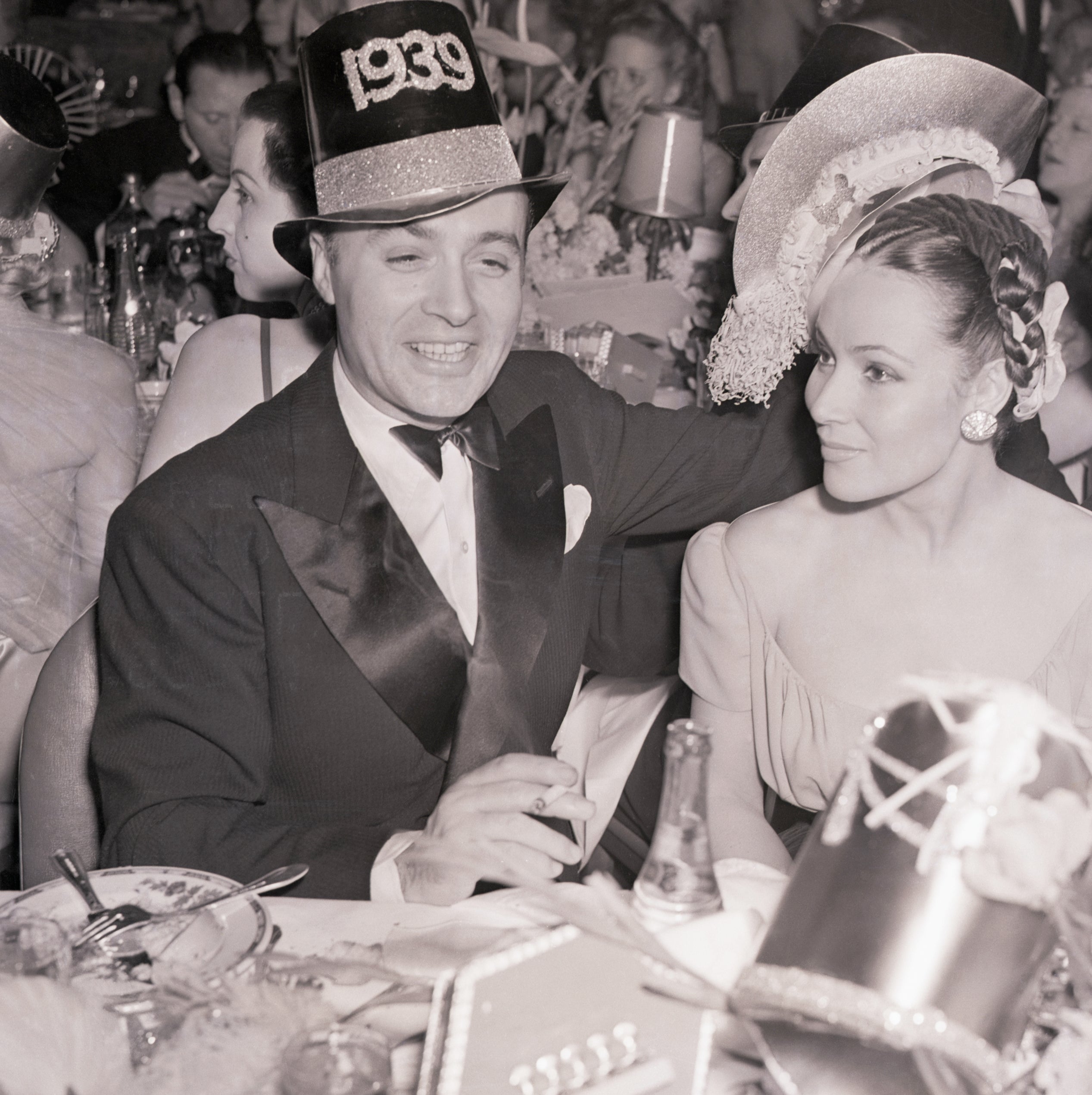 Charles Boyar and Dolores Del Rio at a 1939 New Year's Eve party, wearing formal attire and festive hats, sitting at a table with party decorations