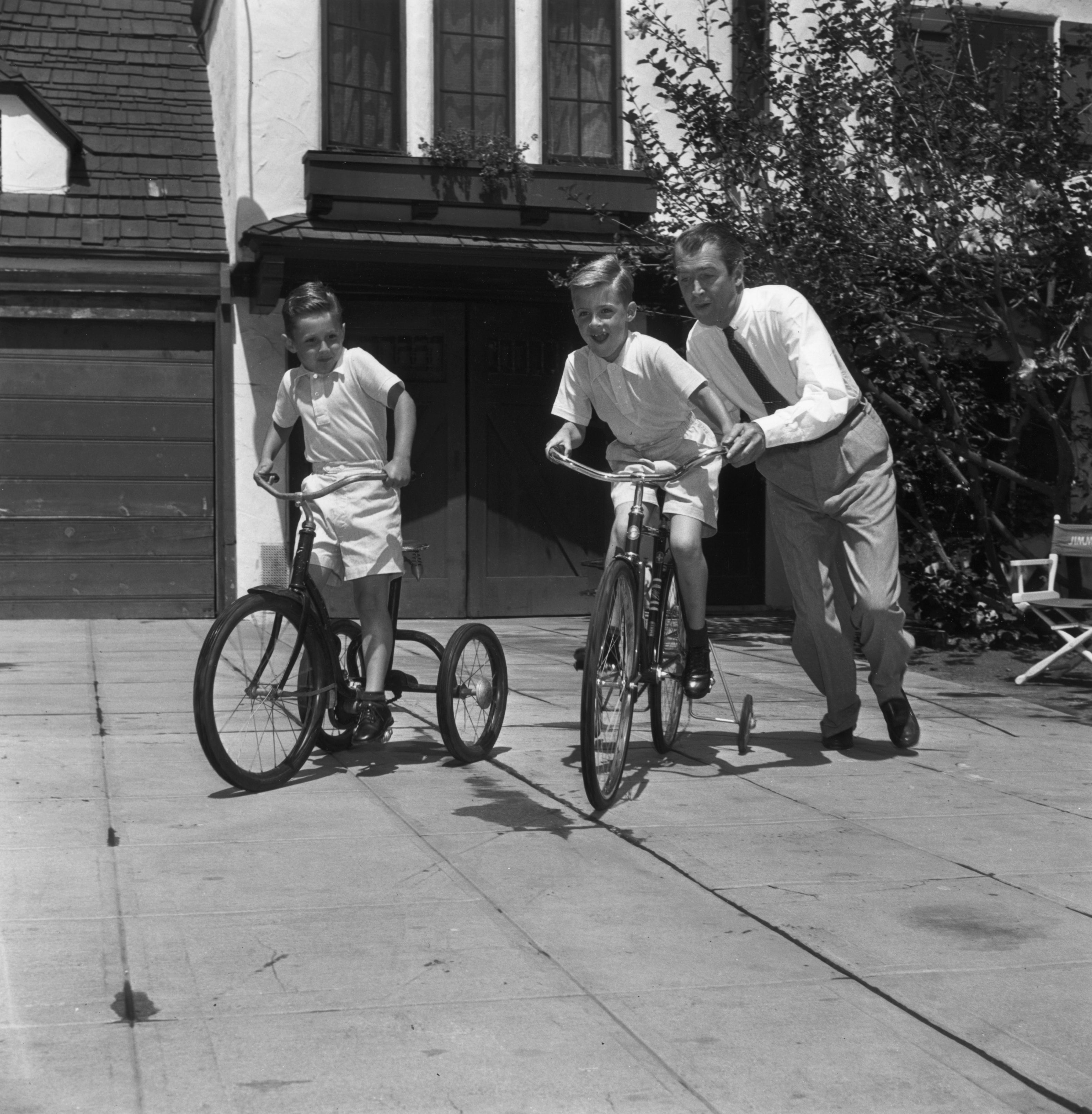 James Stewart helps a child learn to ride a bicycle while another child watches, smiling, on a tricycle in a driveway scene