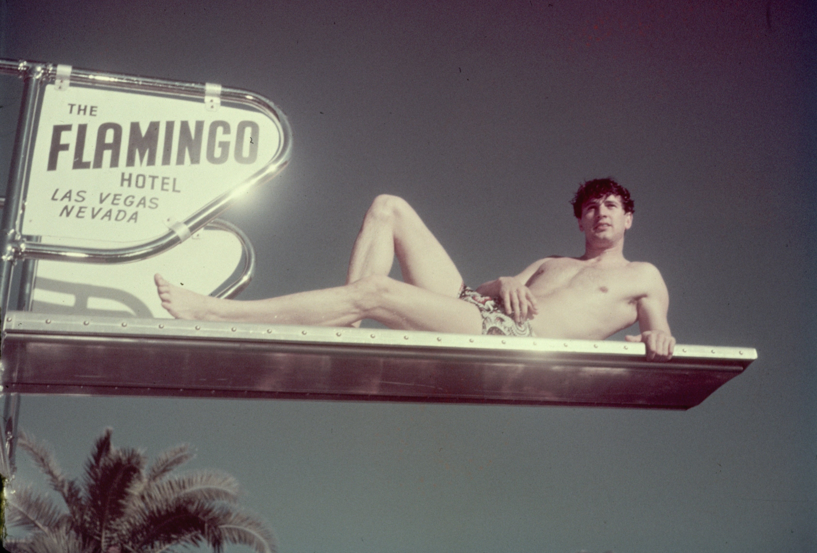 Rock Hudson lounging in a swimsuit on a diving board by a sign for The Flamingo Hotel, Las Vegas, with clear sky background