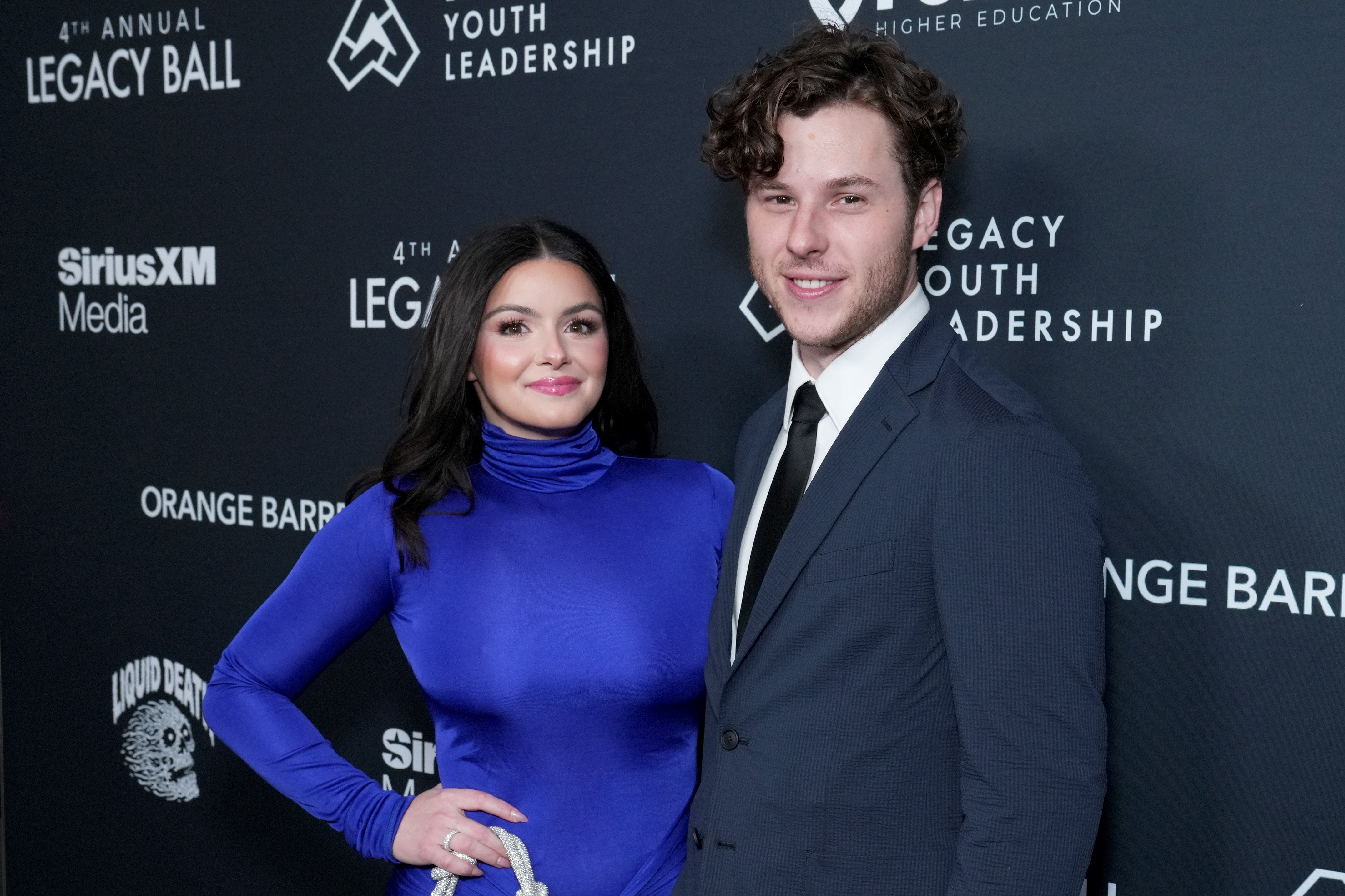 Two people pose at a red carpet event. The person on the left is in a sleek, high-neck dress and the person on the right is in a classic suit