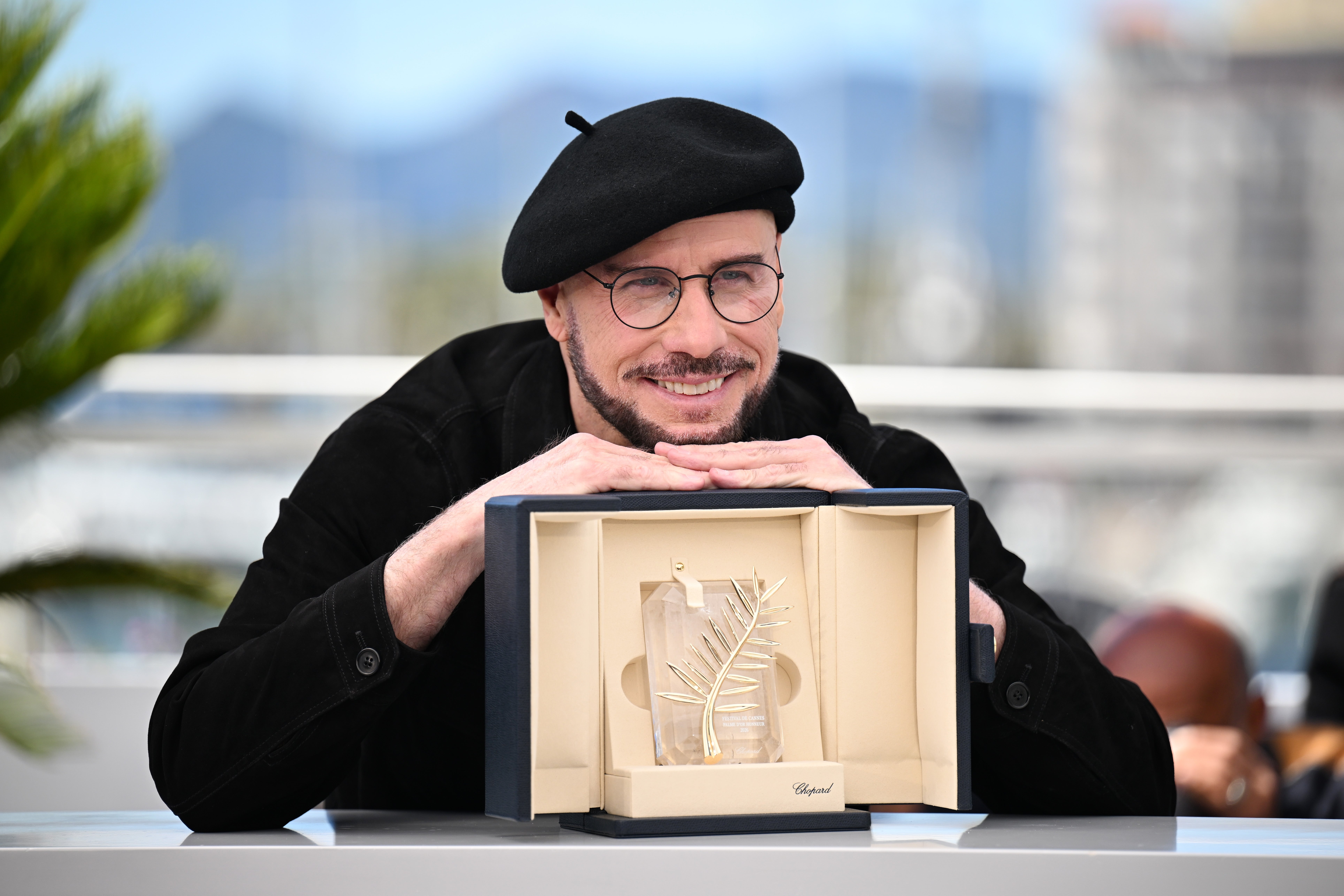 Person in a beret smiling while leaning over an open award box containing a trophy with a palm leaf design
