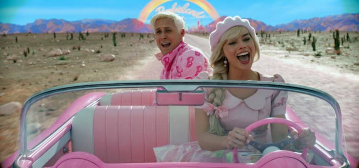 Two people in a pink convertible car, smiling joyfully in a desert setting with a rainbow design in the background