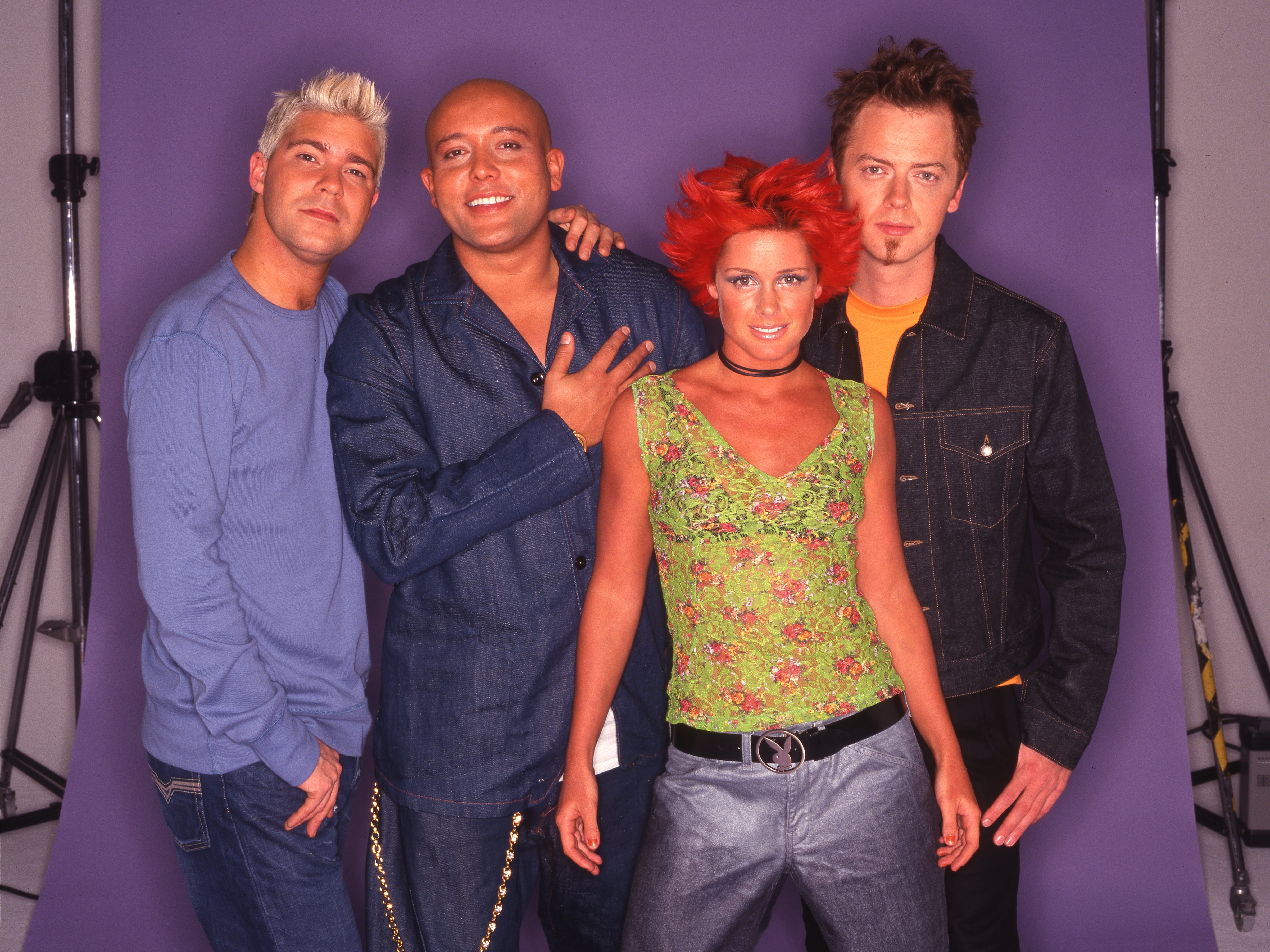 Four people in casual 2000s fashion pose together. One wears denim, another a green floral top. The image has a nostalgic, early 2000s vibe
