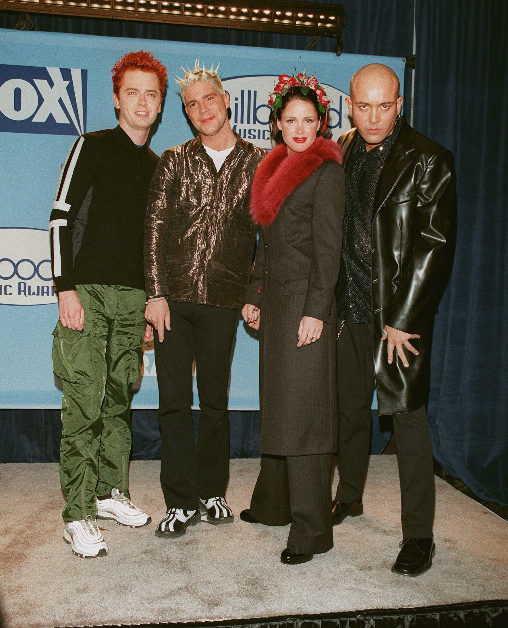 Four people pose on the red carpet, each wearing unique outfits with eye-catching styles, including a leather coat, sparkly tops, and flower headwear