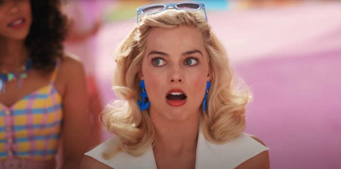 Person with blonde hair, wearing retro-style clothing and bold earrings, looks surprised. Background shows another person with a colorful outfit