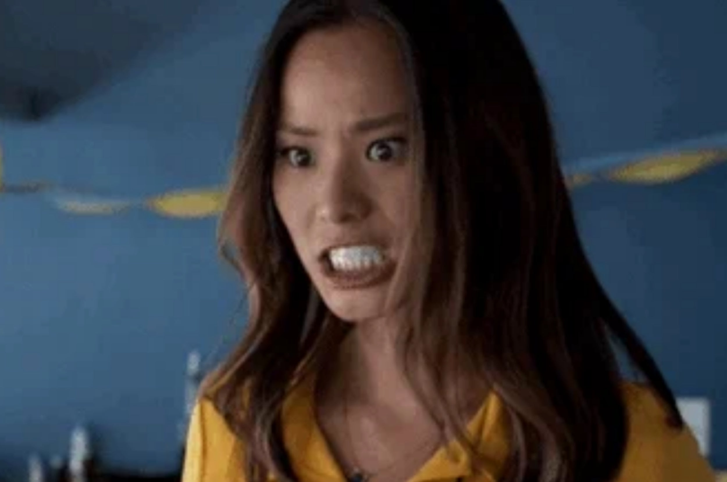 Person with long hair appears frustrated, gritting teeth, wearing a yellow top. Background has blurred party decor