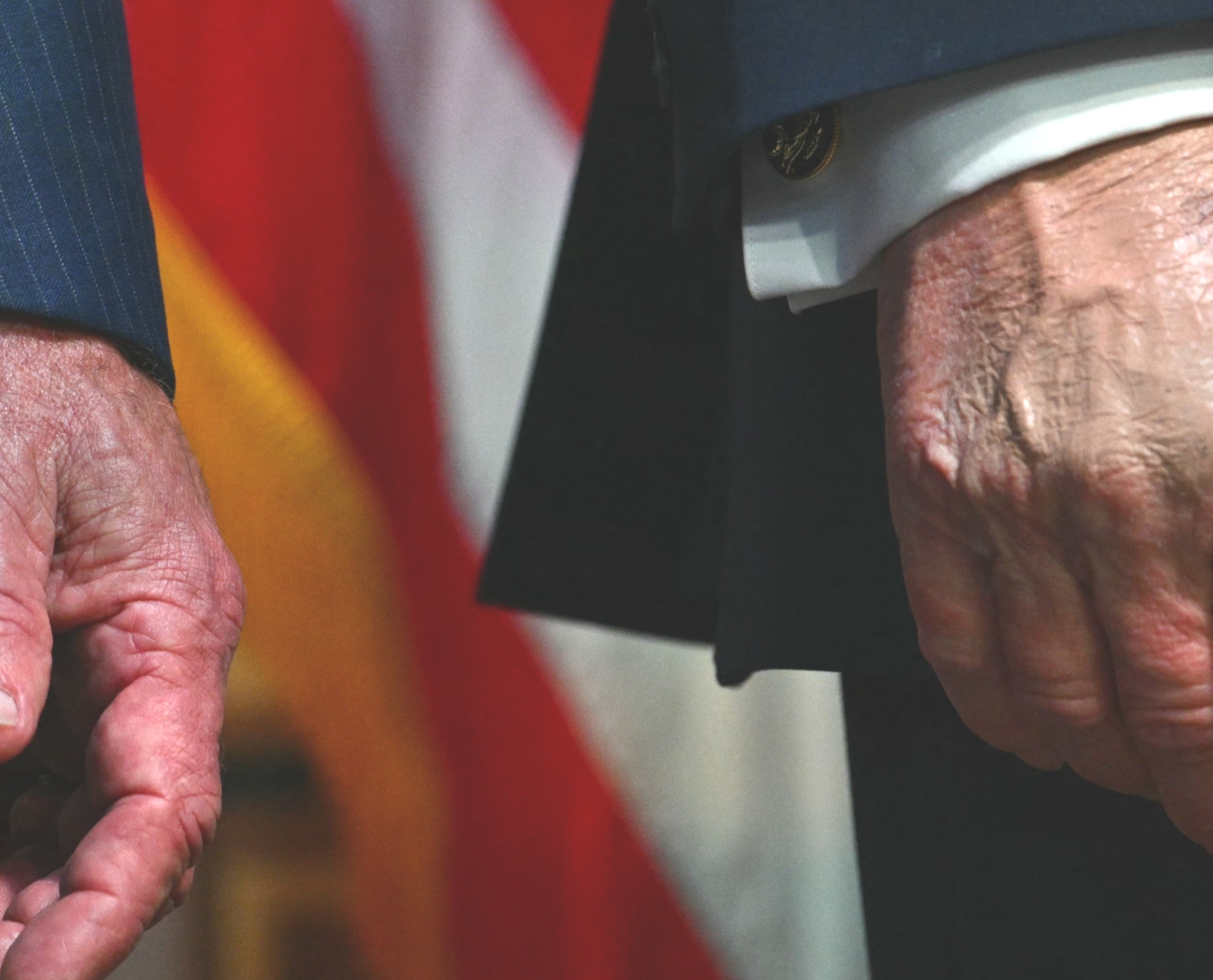 A dark red, hairy, and bruised hand sticks out from a striped suit jacket in front of an American flag background