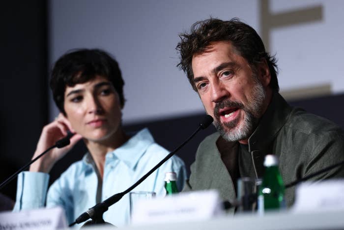 Two people at a press conference, one speaking animatedly into a microphone, wearing a suit jacket. The other looks on attentively