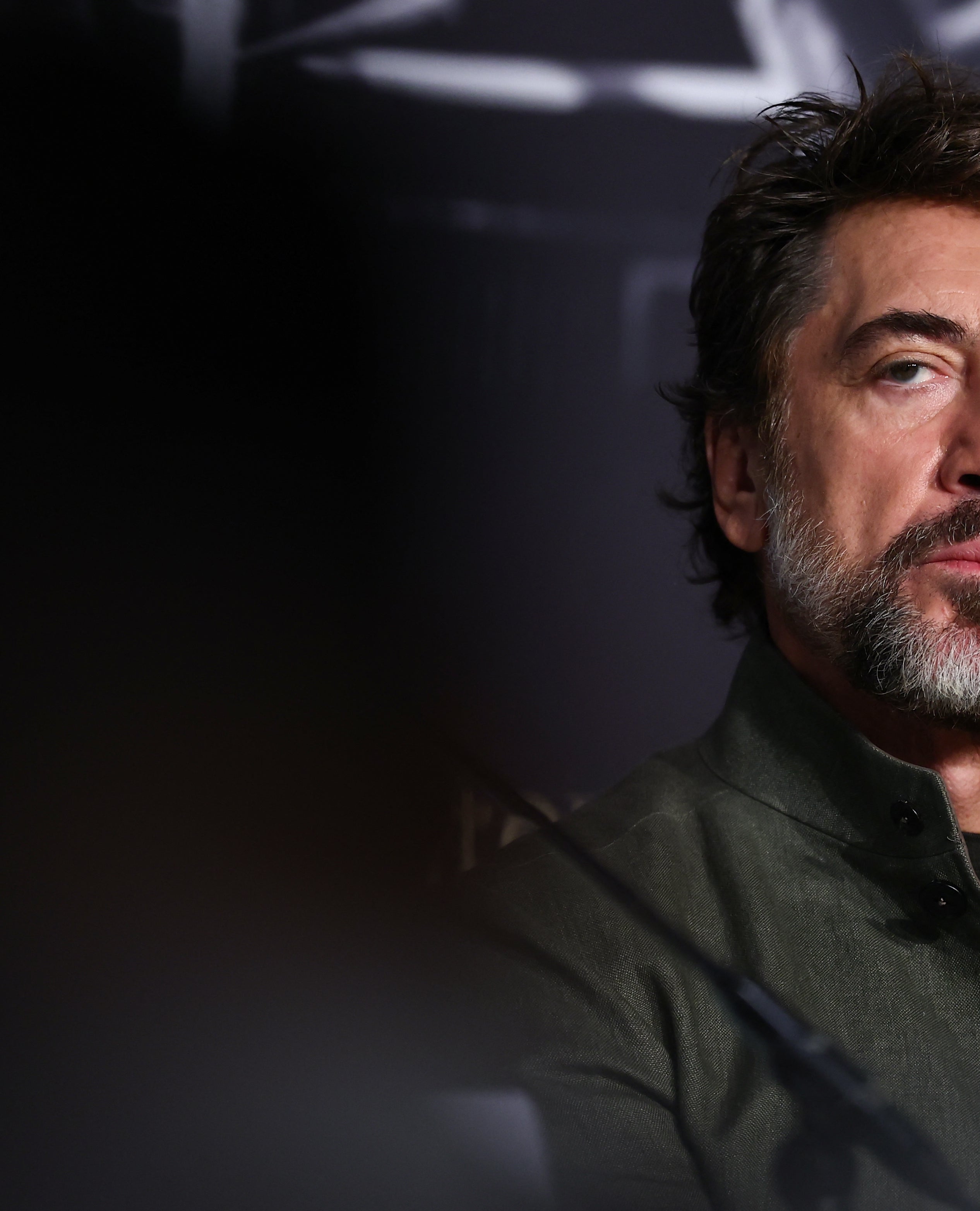 A man with short hair and a beard is at a formal event, wearing a suit jacket and shirt, looking intently forward with a serious expression