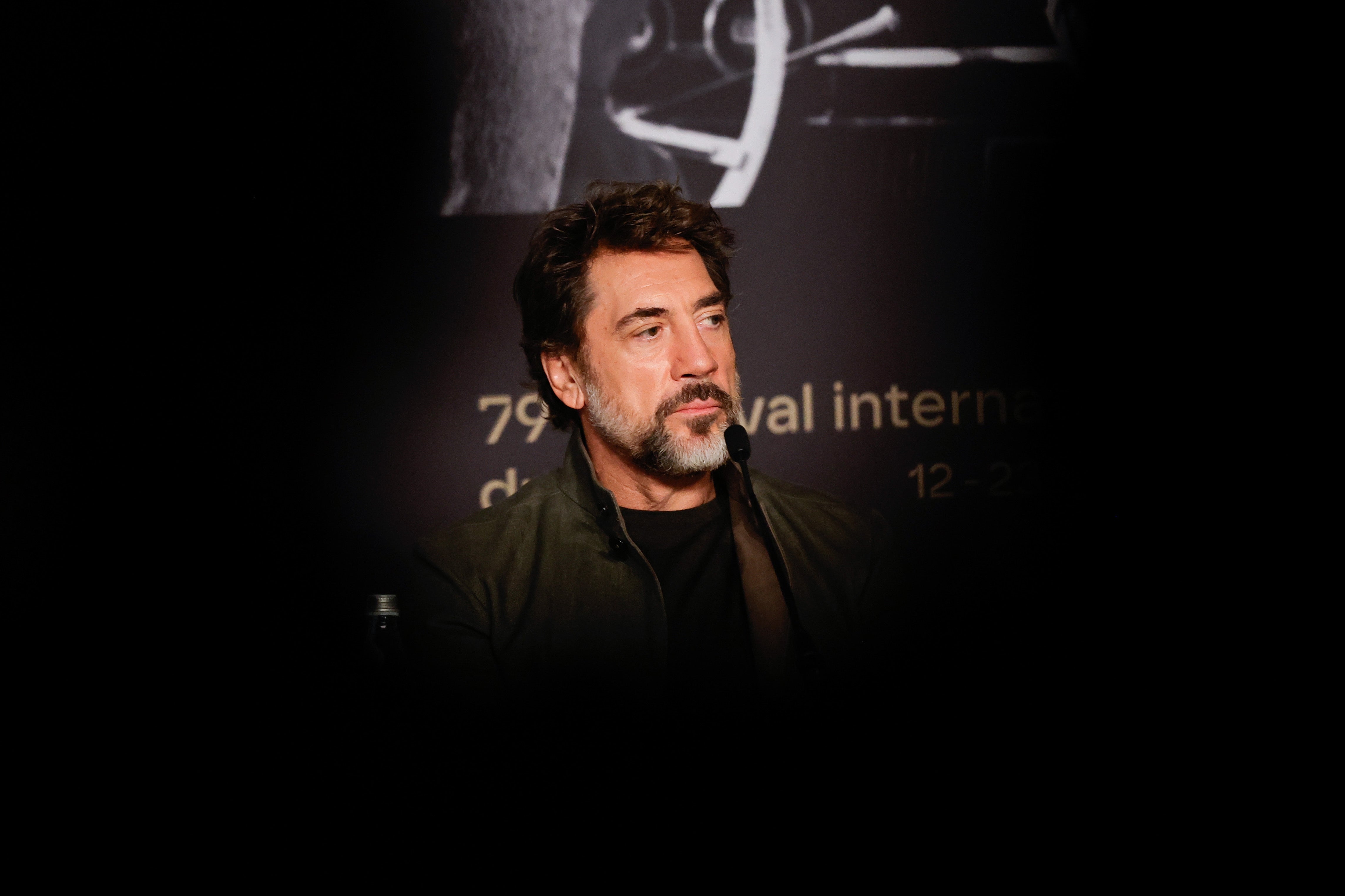 A man with a beard and mustache sits attentively at an event with text behind him