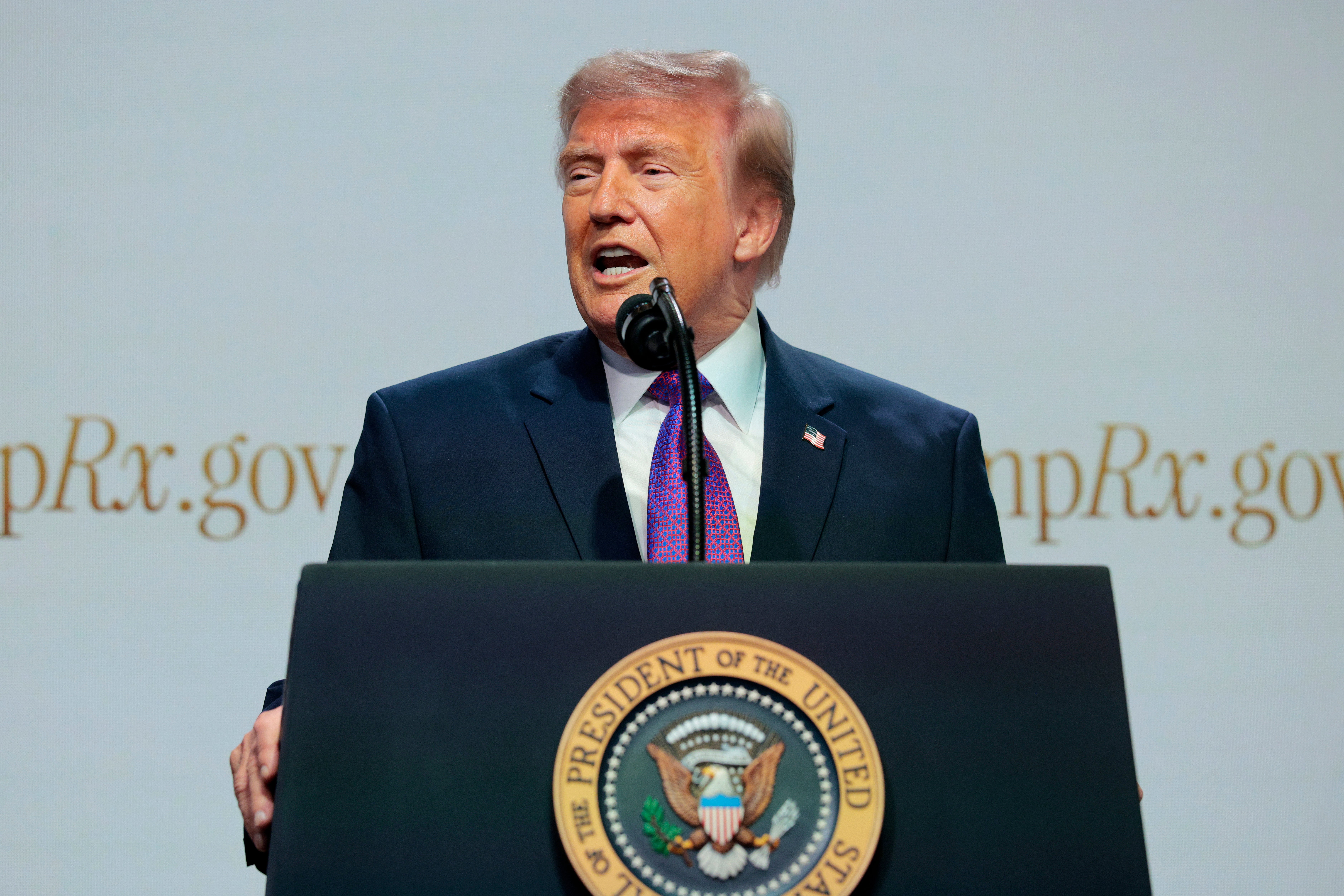 Donald Trump is speaking at a podium with the Presidential Seal, in front of a backdrop featuring the text "mpRx.gov."