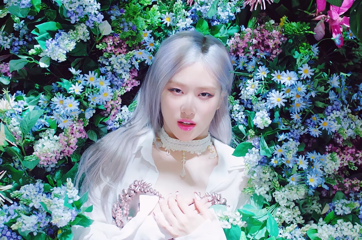 Rosé from Blackpink in "How You Like That" music video with long hair in a white top, surrounded by a rich array of flowers and greenery, looking directly at the camera