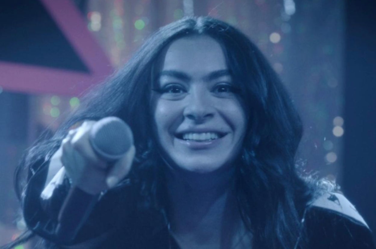 A person with long hair smiles broadly while holding a microphone, as if engaging with an audience