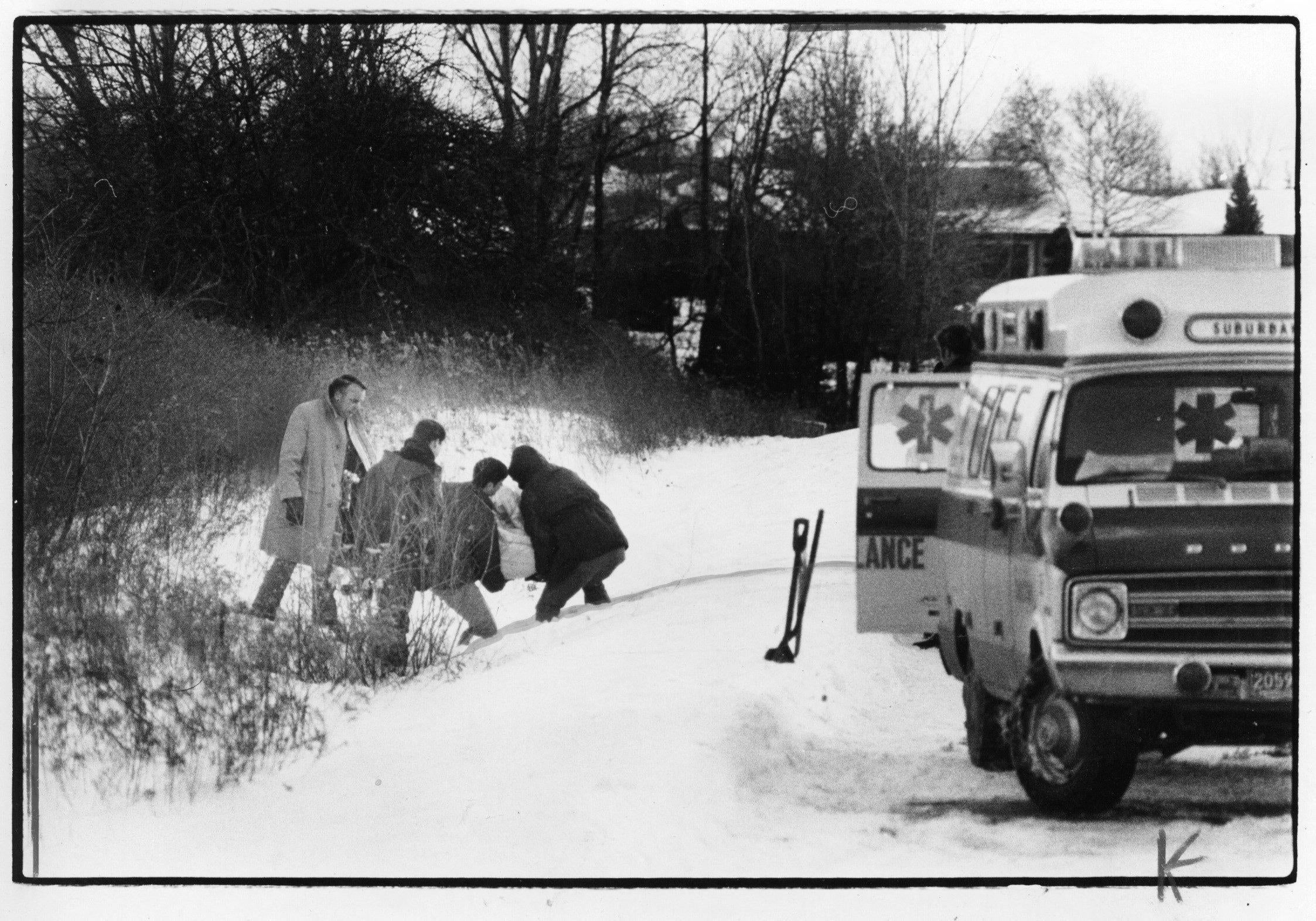 People assisting someone on a snowy path next to an ambulance