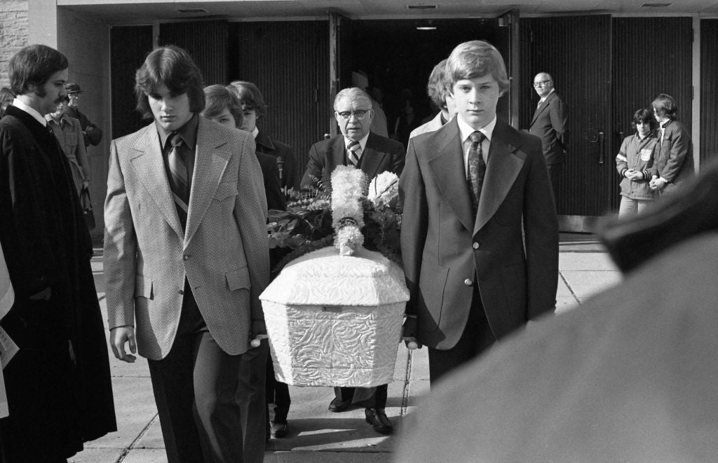 People carrying a floral-covered casket at a funeral, exiting a building. The mood is solemn and the crowd watches respectfully