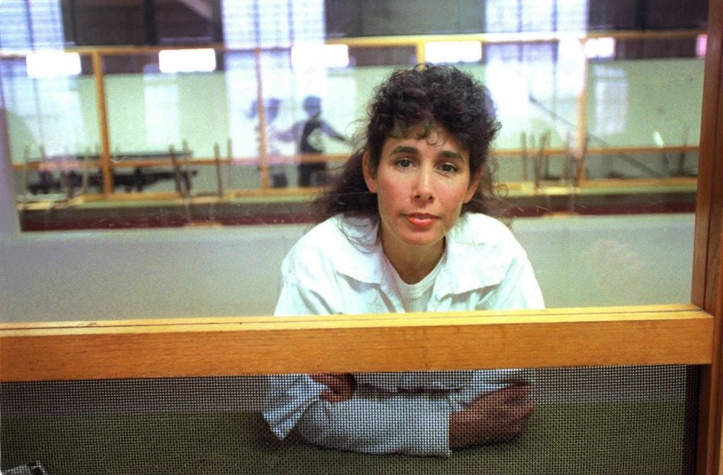 A person with curly hair, wearing a white lab coat, leans on a counter behind a mesh screen in an industrial setting