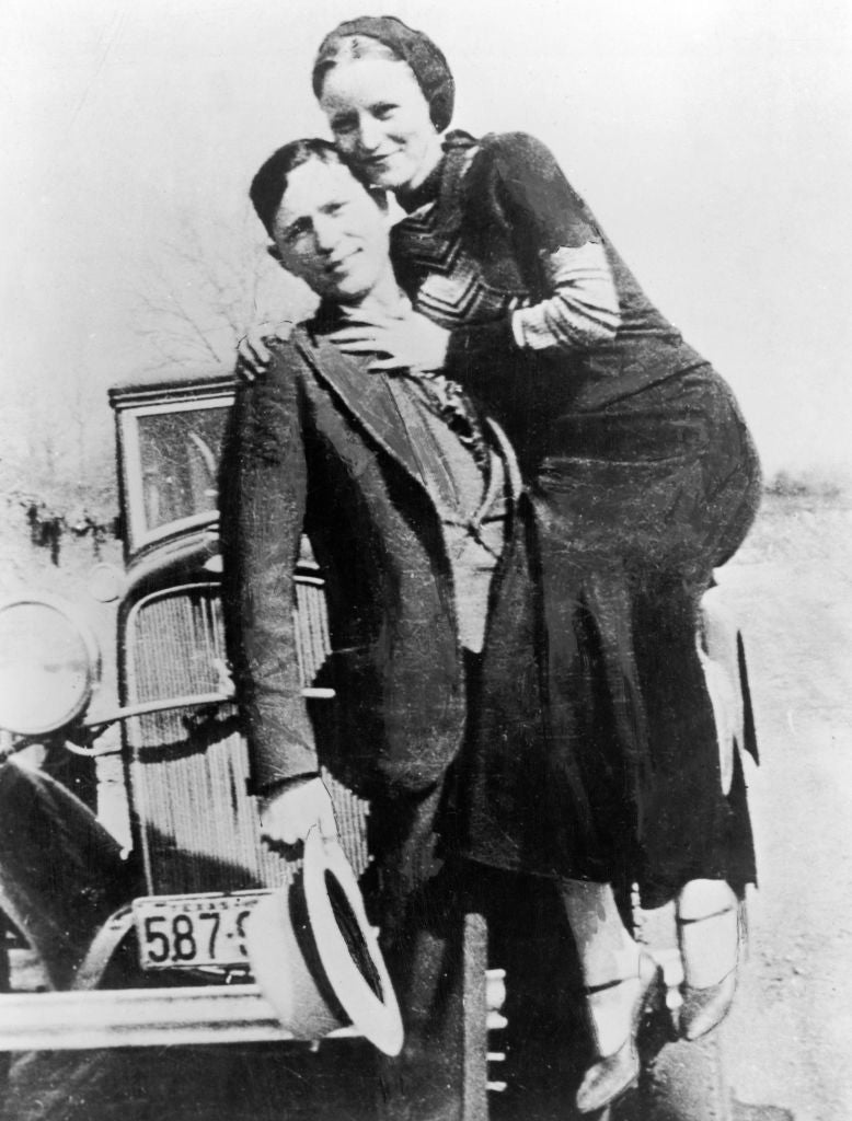 Two people pose playfully on a car in vintage attire, with one sitting on the other's lap, reflecting a candid and lighthearted moment
