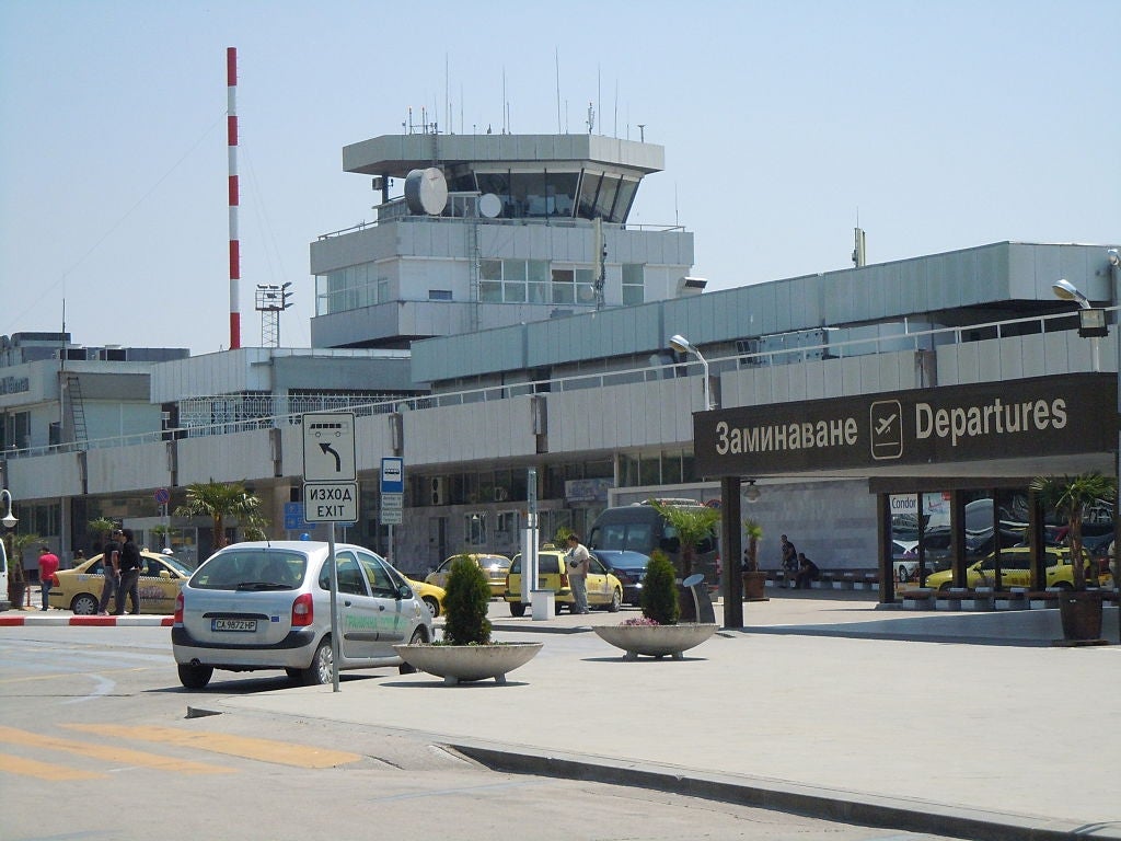 Airport terminal exterior with visible control tower, taxi queue, and parked cars. Signs indicate directions for departures and exits