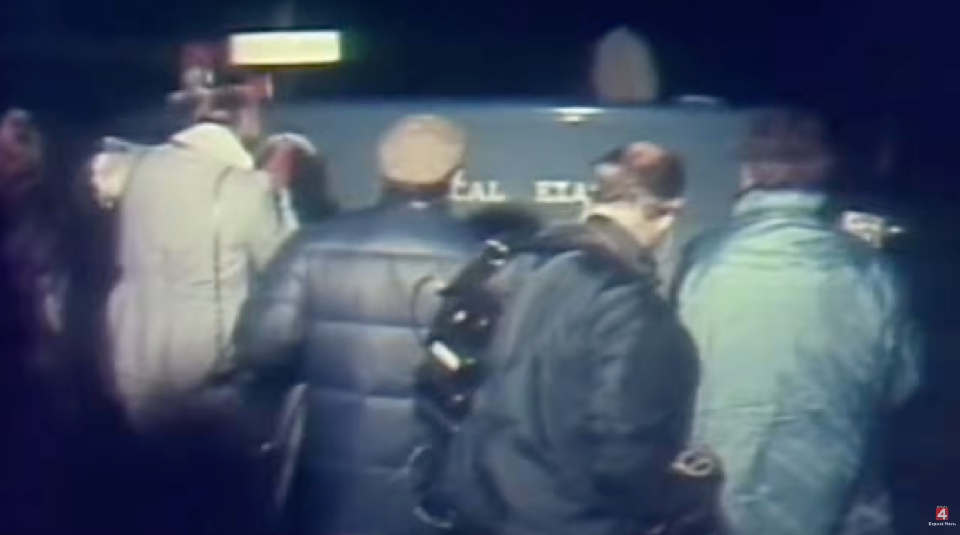 A crowd of people, some with cameras, gather around a vehicle at night, possibly capturing a news event or scene