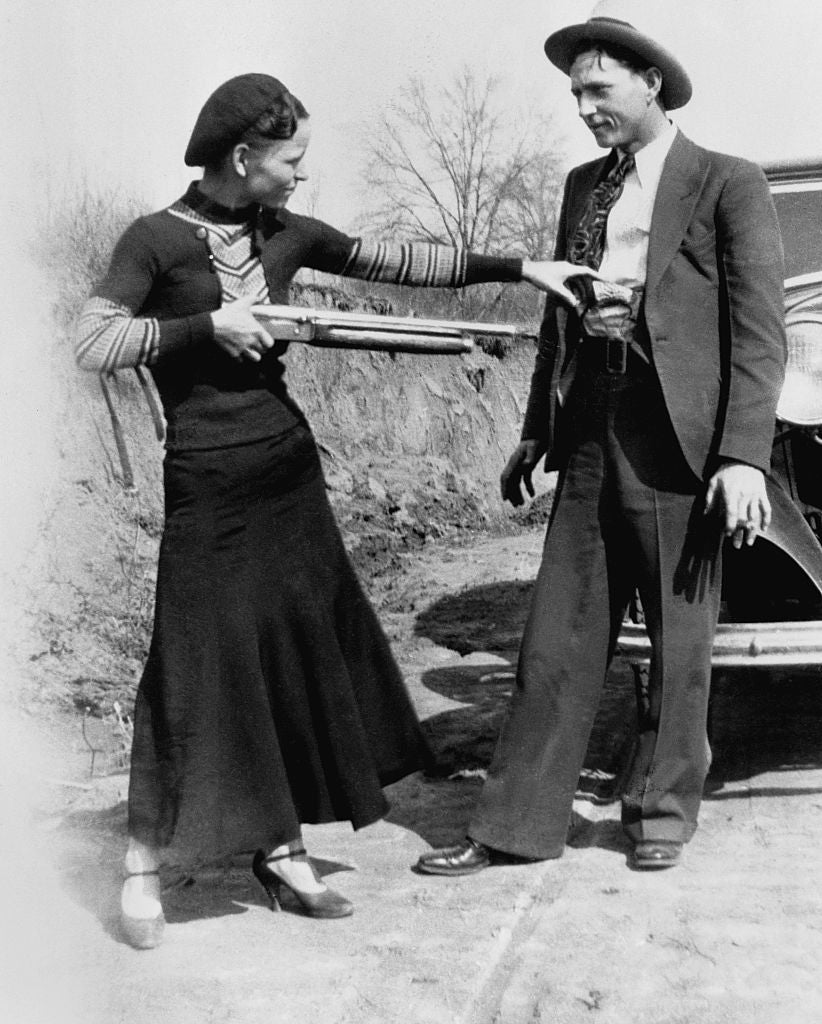 A woman in a beret and a long dress playfully points a shotgun at a man in a suit and hat near a car