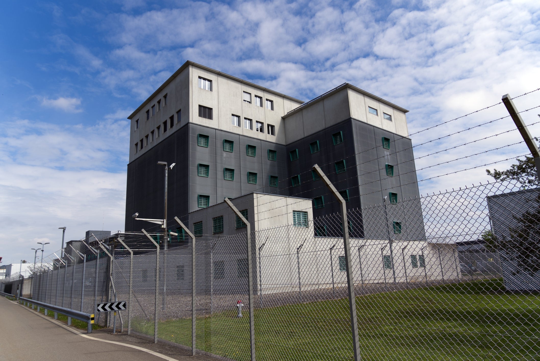 A modern prison building with high walls and barbed wire fences, viewed from the road with a clear sky above