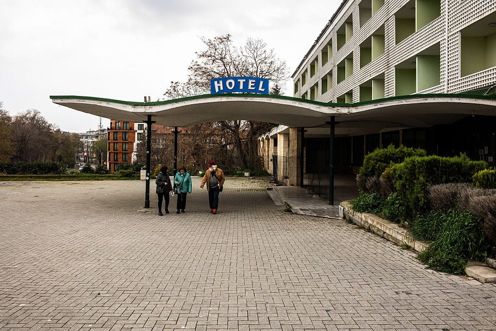 Three people walk under an old hotel entrance with a retro sign, surrounded by greenery and an unoccupied courtyard